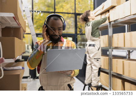Black male staff enjoying depot work with music on headphones, handling operations in a small-scale distribution facility. Preparing parcels for online shopping delivery and product handling. Black male staff enjoying depot work with music on headphones, handling operations in a small-scale distribution facility. Preparing parcels for online shopping delivery and product handling. 133565733