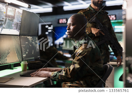 Army unit working in governmental facility operating as surveillance monitoring center filled with digital equipment. African american man doing job in base of operations Army unit working in governmental facility operating as surveillance monitoring center filled with digital equipment. African american man doing job in base of operations 133565761