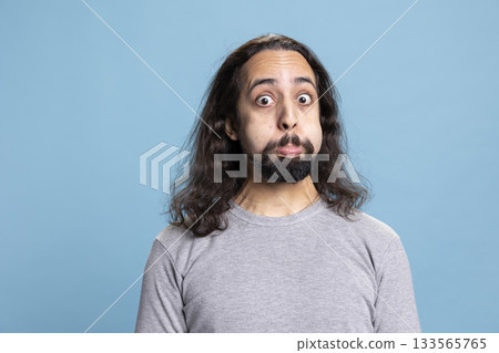 Playful young man with a long hair and beard making a goofy face in the studio. Carefree guy radiates humor and joy, capturing a lighthearted moment full of laughter and casual personality. 133565765