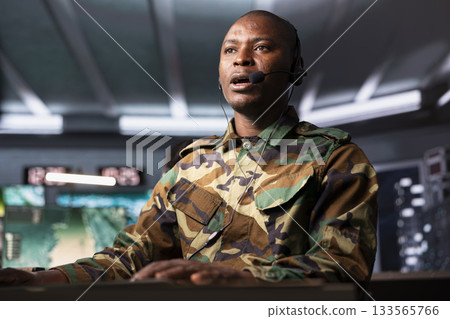 Military dispatcher in base of operations ready to respond to emergencies and communicate with soldiers on front lines. African american telecommunications operator in uniform talking in headset mic Military dispatcher in base of operations ready to respond to emergencies and communicate with soldiers on front lines. African american telecommunications operator in uniform talking in headset mic 133565766