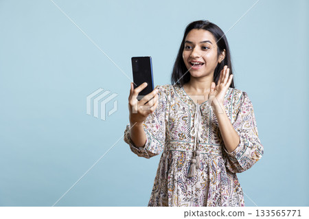 Smiling woman having friendly conversations during teleconference meeting using smartphone, studio background. Indian person having fun catching up with friends during web video call. 133565771