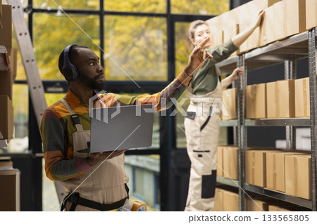 E-shop employee listens to favorite music on headset and counting packages, overseeing quality control and inventory check with laptop. E-business shipments with in house logistics. 133565805