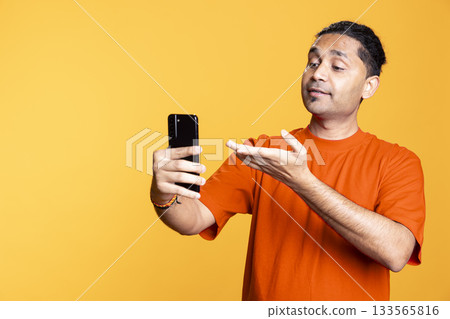 Joyous Indian man greeting people over online videocall on his phone, doing friendly gesture and chatting with friend. Excited young man bonding with someone using mobile phone webcam. 133565816