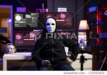 Evil programmer concealing identity using phone to film ransom video in secret base. Scammer with anonymous mask using smartphone to record blackmail footage, threatening victims 133565820