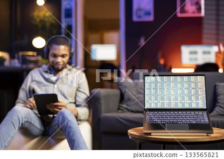 Young Black man relaxes on sofa and uses PC to track forex trades, analyzing stock market graphs for financial growth. Remote worker reviews income trends and trading data for online investments. 133565821