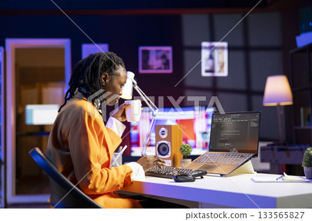 Software developer enjoying cup of coffee and writing code on laptop in apartment office, using Java programming languages. Woman drinking beverage, remotely working on fixing database errors 133565827