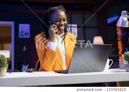 Cheerful teleworker talking in phone call with coworker, using laptop in apartment office to solve work tasks. Smiling teenager doing telephone call with colleague, working together to finish project 133565828