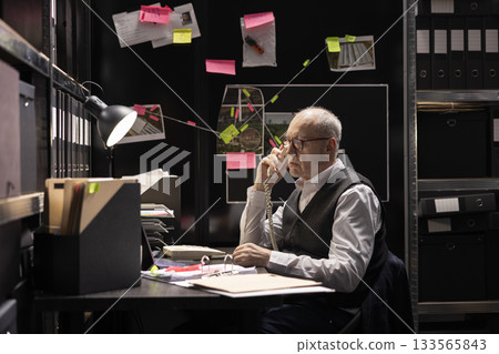 Experienced commissioner uses a landline phone for telecommunication, briefing police department while surrounded by records and evidence in a mission to trace criminal connections. 133565843