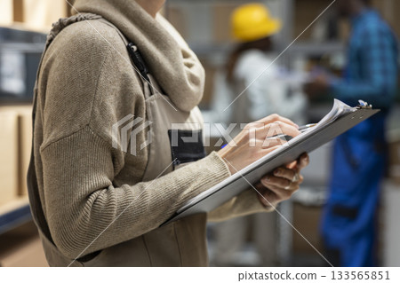 Logistics worker in a depot coordinating storage and package handling, solving distribution tasks before shipment process. Warehouse interior features crates and shelving for supply chain. 133565851