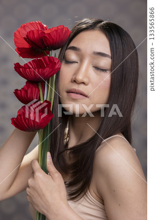Beautiful asian woman model closing her eyes next to red carnations bouquet, posing with glossy smooth skin for a beauty skincare advertisement. Proud feminine pose with natural look. Beautiful asian woman model closing her eyes next to red carnations bouquet, posing with glossy smooth skin for a beauty skincare advertisement. Proud feminine pose with natural look. 133565866