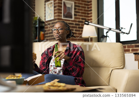 African american woman sitting comfortably on couch, enjoying comedy series with burger and beer. Female entrepreneur relaxing at home after busy day, watching TV for leisure and entertainment. 133565894