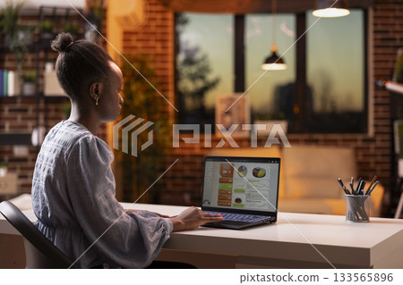 Modern black woman working remotely on business reports and presentations at desk. Confident female entrepreneur using laptop, focusing on online tasks and analyzing company sales revenue at home. 133565896