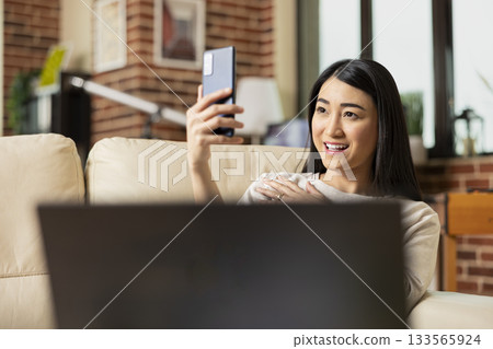 Happy businesswoman relaxes on sofa, taking selfie with smartphone. Cheerful female freelancer captures moment for social media while enjoying downtime at home with mobile device in hand. Happy businesswoman relaxes on sofa, taking selfie with smartphone. Cheerful female freelancer captures moment for social media while enjoying downtime at home with mobile device in hand. 133565924