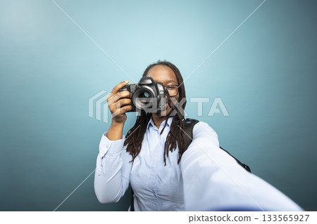 POV of Black female photographer recording herself with a DSLR. African American woman in blue shirt, smiling while documenting her photography journey in a studio with confidence. 133565927