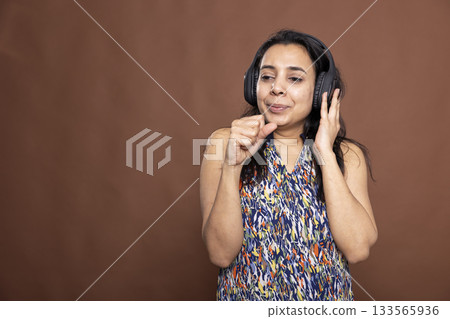 Young female adult wearing wireless headphones, raising her fist to mouth as if singing along. Indian model fully immersed in music, enjoying the audio experience and using her hand like microphone. Young female adult wearing wireless headphones, raising her fist to mouth as if singing along. Indian model fully immersed in music, enjoying the audio experience and using her hand like microphone. 133565936