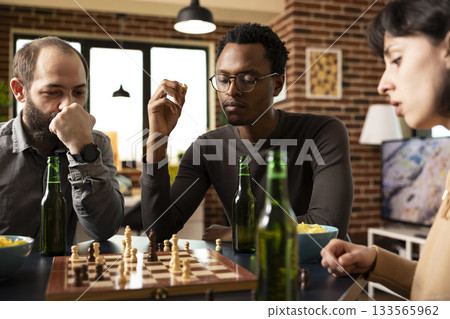 Multiethnic group of friends seated around table, enjoying a board game with snacks. African american man with chess piece in hand, looking at chessboard on table along with his caucasian companions. 133565962