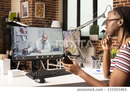 Black woman talking to doctor via desktop video call, seated at home desk with pills bottle in hand. African American patient discussing prescription medicine with physician through computer screen. 133565965