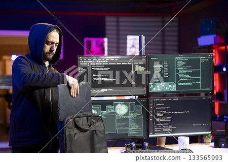 Hacker packing laptop after finishing coding stealth malware to evade security. Evil man putting notebook in backpack after finishing work on script hiding malicious code from detection systems 133565993