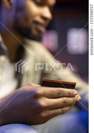 African american man entering banking credentials for online shopping, sitting on the couch to purchase during sale season. Young adult transferring money to pay for his web orders. 133566057
