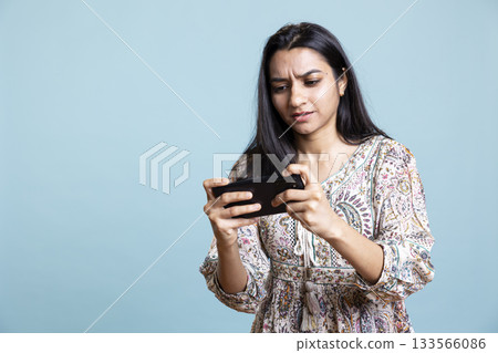 Happy indian woman entertained by videogames on smartphone, enjoying leisure time. Gamer enjoying game on phone, having fun defeating enemies, standing against studio background. Happy indian woman entertained by videogames on smartphone, enjoying leisure time. Gamer enjoying game on phone, having fun defeating enemies, standing against studio background. 133566086