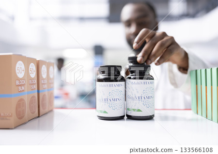 Black male pharmacist stocking medicine and checking pill supplies at the pharmacy shop, ensuring proper arrangement and packaging of health products for safe storage and easy access. 133566108