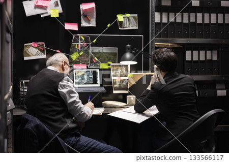 Two inspectors conducting detective work using a landline phone for inquiry and reporting in an incident room. Detectives cross checking evidence with the forensic department on the line. Two inspectors conducting detective work using a landline phone for inquiry and reporting in an incident room. Detectives cross checking evidence with the forensic department on the line. 133566117