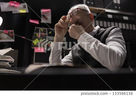 Older private investigator dealing with stress and pressure due to illegal activity, rubbing his eyes and struggling with workload. Agent studying records and classified files in an incident room. 133566138