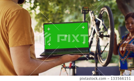 Close-up shot of laptop with isolated chromakey display carried by male cyclist while black woman dismantles damaged bicycle wheel. Couple doing bike maintenance with minicomputer having greenscreen. Close-up shot of laptop with isolated chromakey display carried by male cyclist while black woman dismantles damaged bicycle wheel. Couple doing bike maintenance with minicomputer having greenscreen. 133566151