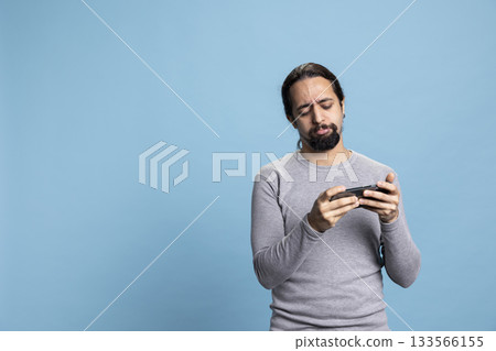 Overwhelmed displeased guy losing a video games championship, feeling dissatisfied and sad about the lost competition. Cyber mobile gaming against blue background in studio. 133566155