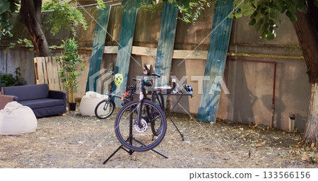 Two bicycles in backyard waiting for repair and maintenance with professional equipment as annual summer activity. Broken bike positioned on repair-stand for examining and fixing outside. 133566156