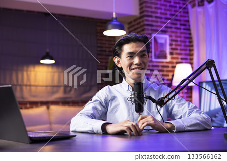Young asian man hosting podcast at home studio desk with microphone and laptop. Male podcaster smiling warmly, expressing enthusiasm while recording digital media content for online platforms. Young asian man hosting podcast at home studio desk with microphone and laptop. Male podcaster smiling warmly, expressing enthusiasm while recording digital media content for online platforms. 133566162