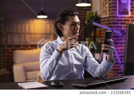 Focused asian man working late at home, using smartphone for video call with colleagues in different time zone. Businessman shares strategy ideas and gestures while discussing project details remotely 133566165