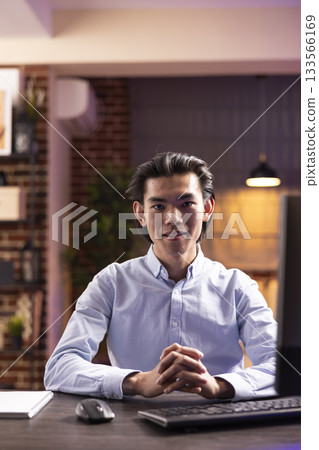 Asian man looks at camera, sits in confidence with computer and notebook for evening online tasks. Male freelancer with clasped hands on table, ready to achieve business goals while working from home. Asian man looks at camera, sits in confidence with computer and notebook for evening online tasks. Male freelancer with clasped hands on table, ready to achieve business goals while working from home. 133566169