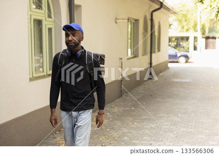 African American courier delivers a hot meal order stopping at doorstep. Delivery man uses the intercom to confirm the delivery location and notify the customer inside, quick service. 133566306