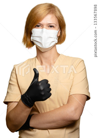 A female doctor, a healthcare worker. Vertical shot. She is making the SUPER sign with one hand. Her thumb is pointing up. She is wearing a beige uniform, a medical protective mask, and black gloves 133567898