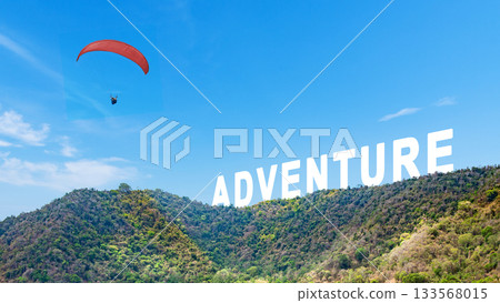 Outdoor scene featuring a paraglider soaring through a clear blue sky with Adventure text over lush green hills. Concept of travel 133568015