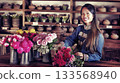 Smiling Asian saleswoman arranging vibrant roses and tulips in ceramic vases in flowers store, looking at the camera. Natural wood and warm lighting. 133568940