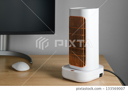 Modern electric heater on a wooden desk beside a computer monitor 133569007