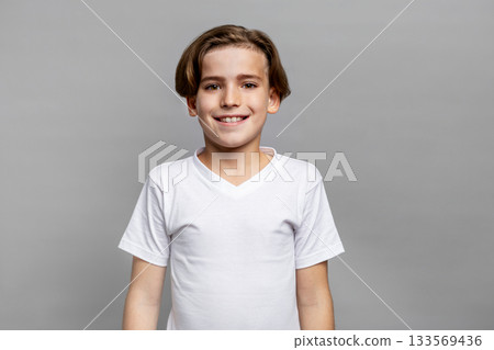 Smiling brunette boy with brown eyes posing for studio portrait, looking at camera in white t-shirt, isolated on neutral gray background, expressing happiness and positive emotion 133569436