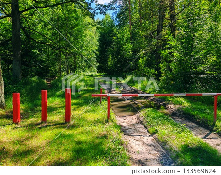Red and White Barrier Blocking Forest Path. metal barrier across a muddy dirt road leading into a dense green summer forest. Restricted access, nature protection and outdoor recreation concept. Red and White Barrier Blocking Forest Path. metal barrier across a muddy dirt road leading into a dense green summer forest. Restricted access, nature protection and outdoor recreation concept. 133569624