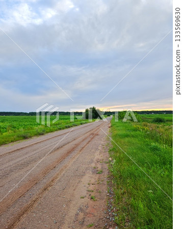 Endless Rural Dirt Road at Sunset. Long straight dirt road stretching through vast green fields and wildflowers under dramatic cloudy sky at golden sunset Endless Rural Dirt Road at Sunset. Long straight dirt road stretching through vast green fields and wildflowers under dramatic cloudy sky at golden sunset 133569630