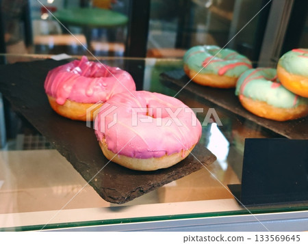 Pink Glazed Donuts in Bakery Display Case. Freshly glazed pink and mint donuts with colorful icing displayed on black slate trays behind glass in a cozy bakery showcase. 133569645