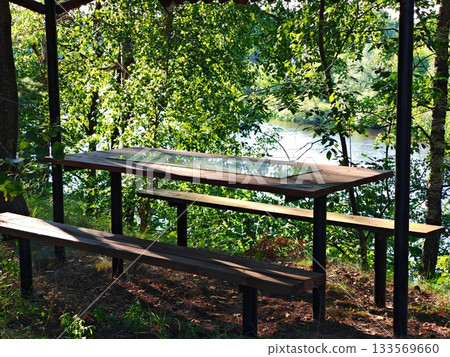 Wooden Picnic Table Overlooking Calm River. Empty rustic wooden picnic table and benches under a simple shelter with a peaceful view of a calm river surrounded by dense green forest. 133569660
