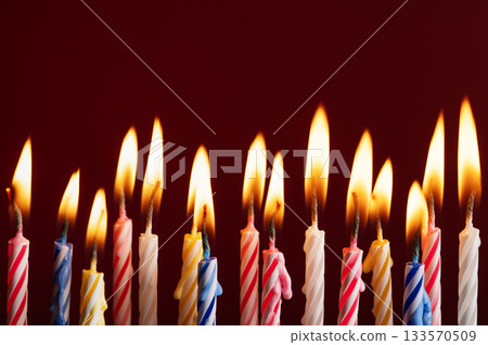 Row of colorful burning birthday candles on red background. Bright flames and vibrant colors with space for text or graphic elements. Ideal for celebration concepts. Row of colorful burning birthday candles on red background. Bright flames and vibrant colors with space for text or graphic elements. Ideal for celebration concepts. 133570509
