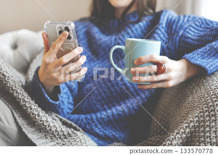 Woman shopping online at home, holding smartphone and warm cup in knitted blanket and sweater, enjoying comfort, convenience and modern digital lifestyle indoors. Cozy winter moment Woman shopping online at home, holding smartphone and warm cup in knitted blanket and sweater, enjoying comfort, convenience and modern digital lifestyle indoors. Cozy winter moment 133570778