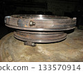 Heavy, industrial component, possibly part of machinery, rests on a textured surface in a dimly lit workshop. The metal shows signs of wear, rust, and bolted connections. Generator seal. 133570914