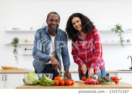 A smiling couple prepares a meal together in a bright, modern kitchen, chopping vegetables. 133571167