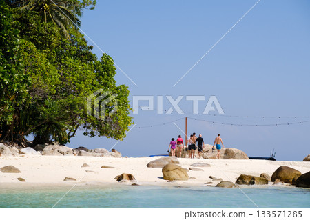 A group of friends is walking on a sunny day on a sandy beach with lush green trees and a clear blue sky by the water. Lang Tengah Island. Malaysia 133571285