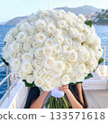 Woman holding massive white rose bouquet, standing on yacht deck with ocean and coastal landscape stretching behind her 133571618