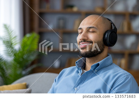 Happy bald bearded man wearing wireless headphones, eyes closed and smiling while listening to music at home, relaxed and peaceful during a quiet moment of leisure Happy bald bearded man wearing wireless headphones, eyes closed and smiling while listening to music at home, relaxed and peaceful during a quiet moment of leisure 133571683